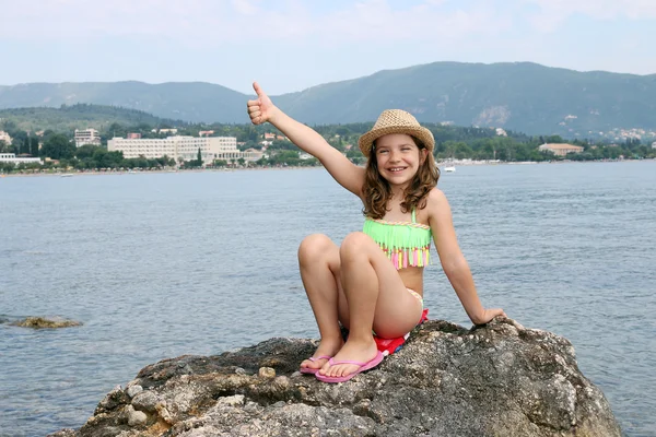 Felice bambina con il pollice alzato seduta su una roccia in riva al mare — Foto Stock