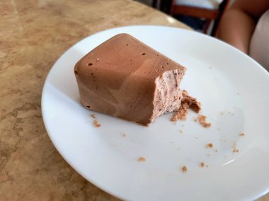 Kare şeklinde zengin, koyu kahverengi çikolatalı bir tatlıya yakın plan, sıklıkla Blok Czekoladowy (çikolata bloğu) veya çikolata şekerlemesi olarak adlandırılır. Tatlının yoğun, pürüzsüz ve hafif gözenekli bir dokusu var. Beyaz tabakta görünen kırıntılar...