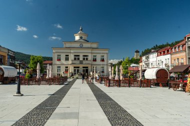 Muszyna, Küçük Polonya Voyvoda, Polonya - 05 Ağustos 2025 - Pazar Meydanı 'ndaki tarihi belediye binasının merkezi manzarası, açık bir yaz gökyüzü altında kafe bahçeleriyle çevrili.