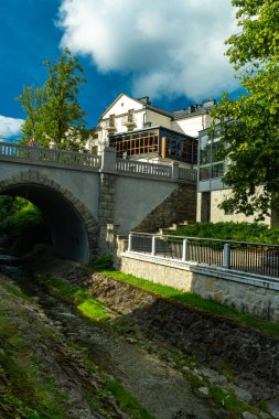 Krynica-Zdroj, Malopolska Voyvoda, Polonya - 12 Ağustos 2025 - Dikey bileşim taş kemer köprüsüne, binaya çıkan merdivenlere ve aşağıdaki yemyeşil ağaçlarla çevrili düzenlenmiş dere yatağına odaklanıyor.