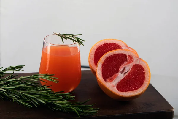 光を背景に木製のテーブルの上に横たわる赤いオレンジ ローズマリーの小枝とオレンジジュースのガラスの二つの半分 — ストック写真