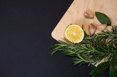 Koyu yapraklar, biberiye filizleri, limon ve karanfil desenleri ile çerçevelenmiş tahta kesme tahtası koyu gri arka planda tasarlanmış.