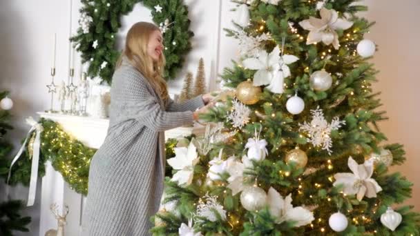 Ευτυχισμένη νεαρή ξανθιά γυναίκα διακόσμηση χριστουγεννιάτικο δέντρο με μπάλα στο σπίτι της — Αρχείο Βίντεο