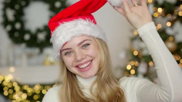 若いですブロンド美しいです笑顔女性でセーターとサンタ帽子オンクリスマスツリー背景 — ストック動画