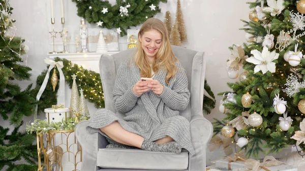 Mosolygó trendi nő szürke hangulatos pulóver ül kényelmes karosszék és szöveges üzenet küldése — Stock Fotó