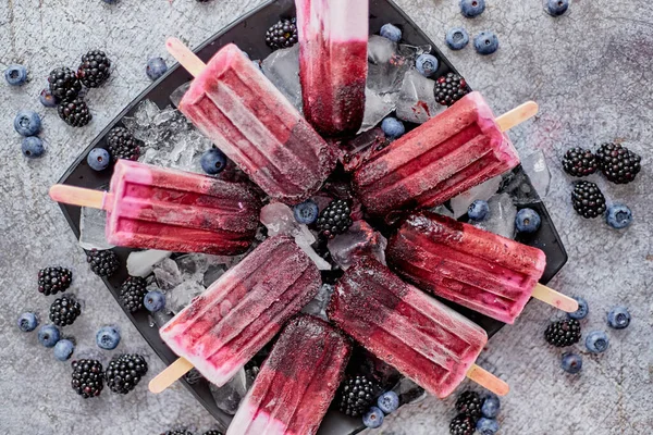 Homemade fresh frozen blueberry and blackberry popsicles on black plate with ice sitting on stone