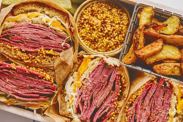 Roti lapis besar dengan daging sapi pastrami dalam kotak kayu. Disajikan dengan kentang panggang, acar — Stok Foto