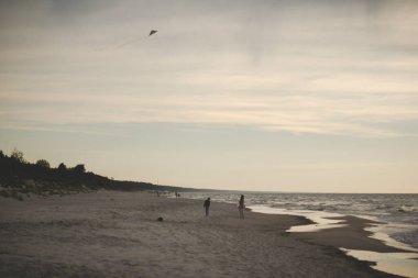 Uçurtmayla yürüyen insanlarla dolu neredeyse boş bir sahil manzarası.