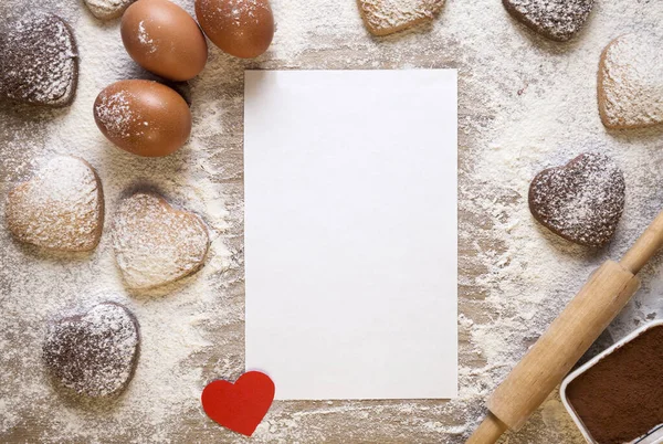 Fond de cuisson avec feuille de papier vierge pour la recette ou le menu, biscuits en forme de coeur, oeufs, farine, rouleau à pâtisserie et petit coeur en papier rouge.. — Photo