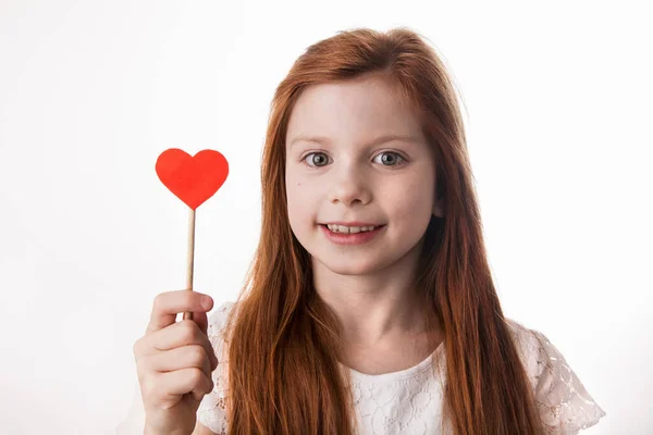 一个红头发的小女孩的画像，手里拿着红心棒。爱、情人节、母亲节、父亲节或快乐生日的概念. 免版税图库照片