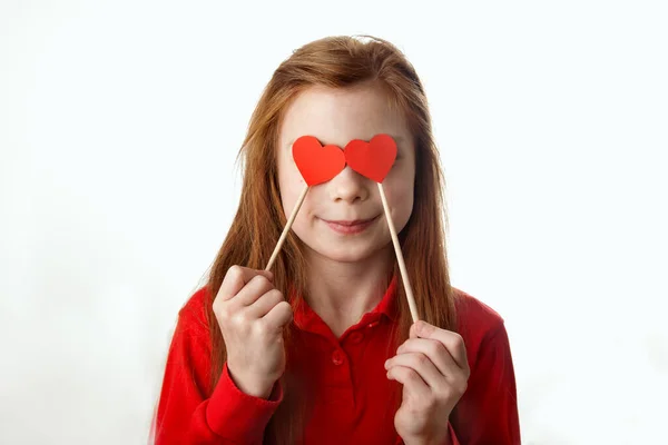Porträt eines kleinen rothaarigen Mädchens, das seine Augen mit roten Herzen bedeckt, lizenzfreie Stockbilder