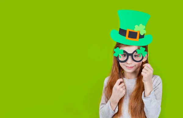 Little smiling girl in green hat of leprechaun and shamrock clover glasses for irish St. Patricks Day on green background —  Fotos de Stock