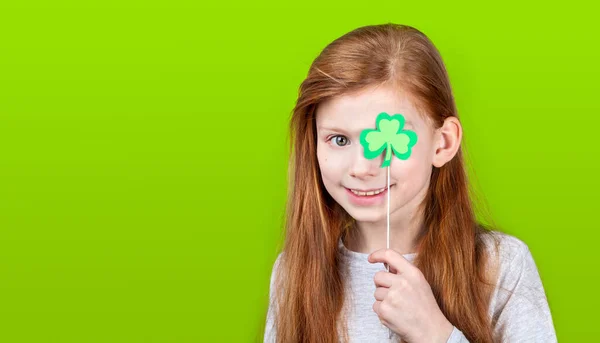 Little smiling caucasian red-haired girl covering her eye with shamrock clover leaf on a stick on green studio background.. — Stockfoto