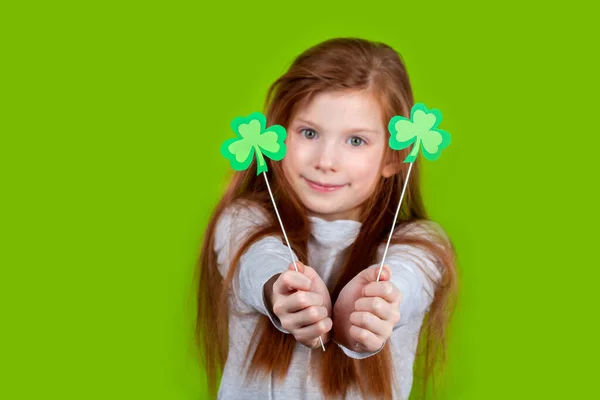Kleines lächelndes kaukasisches rothaariges Mädchen mit zwei Kleeblättern auf Stöcken in der Hand auf grünem Studiohintergrund.. — Stockfoto
