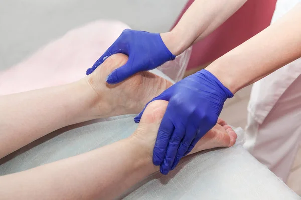 Les mains de masseur en gants de protection bleus touchent les pieds de la patiente faisant une procédure de massage des pieds dans un salon de spa. Photo De Stock