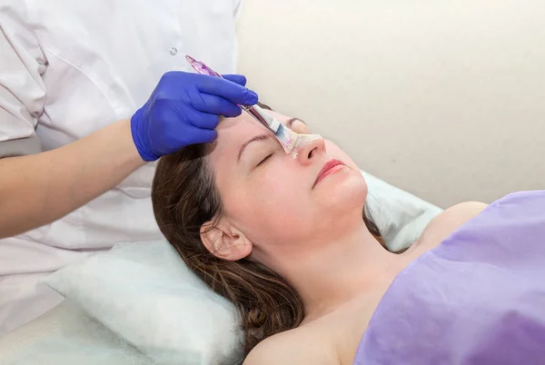 Toepassing van cosmetologie masker op het gezicht van een jonge vrouw in spa salon. Cosmetoloog hand in blauwe beschermende handschoen aanraken patiënt gezicht met borstel.. Rechtenvrije Stockfoto's