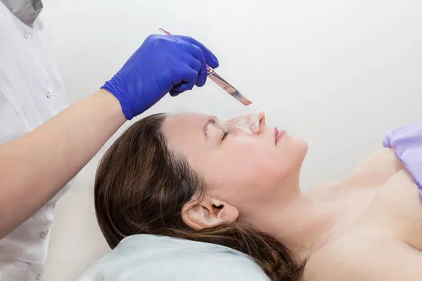 Aplicação de máscara cosmetologia no rosto de uma jovem mulher no salão de spa. Cosmetologist mão em azul luva protetora tocando paciente rosto com escova.. Imagens Royalty-Free