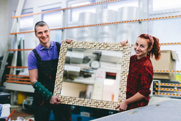 Man and woman showing a mirror with handmade wooden frame in glazier workshop. Small business