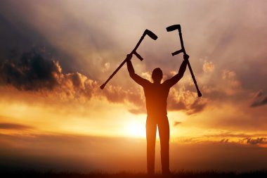 Man raising his crutches at sunset. clipart