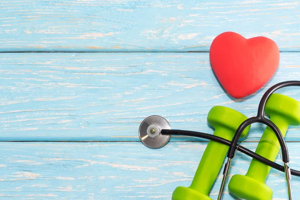 Um coração com um estetoscópio está em um fundo de madeira ao lado de halteres. Coração saudável — Fotografia de Stock