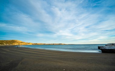 Üzerinde yürüyen bir adamın olduğu bir kumsal. Gökyüzü mavi ve bulutlu. Kumsal kumlu ve su sakin.