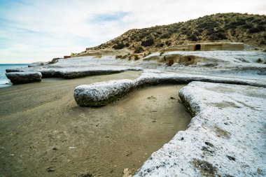 Arka planda su bulunan kayalık bir sahil. Kumsal karla kaplı. Sahilde kayalar var.