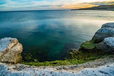 Kenarında kaya duvarı olan bir su kütlesi. Su mavi ve kayalar gri.