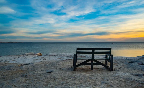 Sahilde bir bank oturuyor. Yedek kulübesi boş. Gökyüzü mavi ve güneş batıyor.