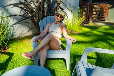 Çimenli bir alanda bir kadın sandalyede oturuyor. Şapka ve güneş gözlüğü takıyor.