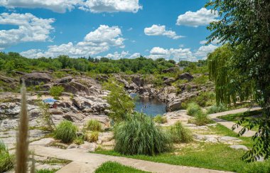 Küçük bir dereyle çimenli bir alana giden bir yol var. Nehir kayalar ve ağaçlarla çevrili.