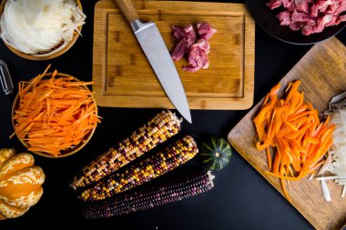 Kesme tahtasındaki et ve bıçak siyah bir masanın üstünde sebzelerle çevrili. 