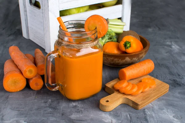 Gri ahşap bir masada şişede taze havuç suyu ve kutuda meyve.