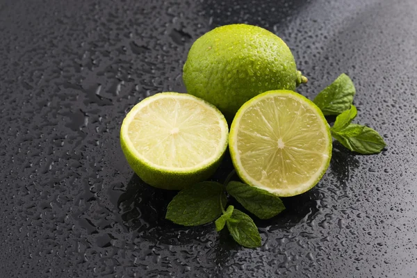 Limas verdes com gotas de hortelã e água sobre fundo preto — Fotografia de Stock