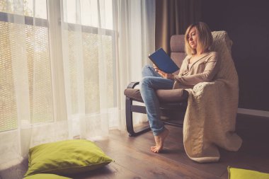 Evde izole edilmiş ve dinlenen genç bir kadın pencerenin yanındaki sandalyede oturmuş kitap okuyor.