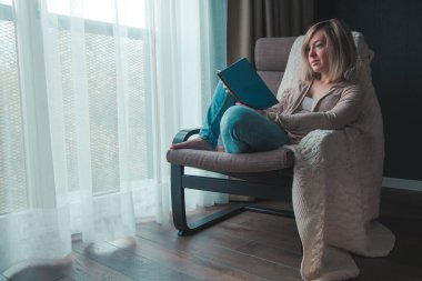 Rahatlatıcı bir kadın evde yalnızken pencerenin yanındaki sandalyede oturmuş kitap okuyor.