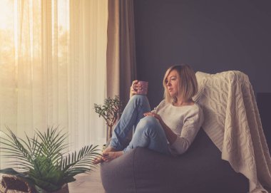 Düşünceli bir kadın kahve içip pencereden dışarı bakarken evdeki fasulye çuvalının üzerinde oturuyor.