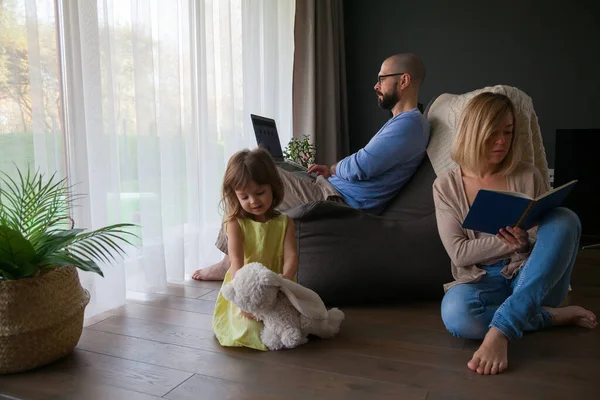 Family spending time together at home - father with laptop, mother reading a book, and their little daughter playing alone with her toy