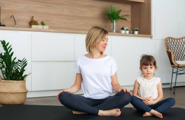 Kadın ve küçük tatlı kızı evde yoga yaparken lotus pozisyonunda oturuyorlar.