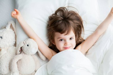 Güzel küçük kız beyaz bir yatakta uyanırken ellerini uzatıyor. Üst görünüm.