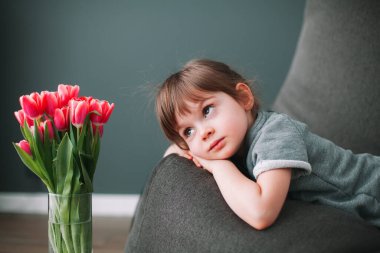 Güzel küçük kız vazodaki pembe lalelere bakıyor, gri fasulye koltuğu üzerinde yatıyor.