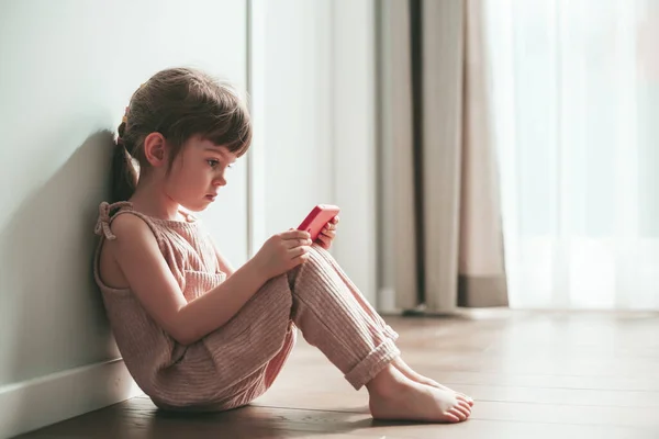 Cute little girl with mobile phone sitting alone on a floor. She is playing games or watching cartoons on smartphone. Digital generation and phone addiction concept.