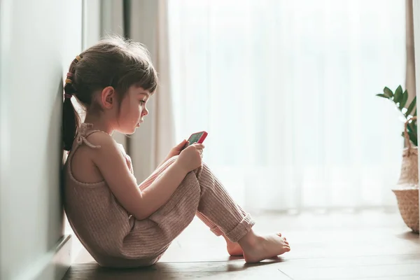 Cute little girl with mobile phone sitting alone on a floor. She is playing games or watching cartoons on smartphone. Digital generation and phone addiction concept.