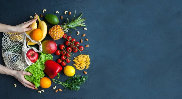 Hands holding a string bag with healthy vegetarian food. Variety of vegetables and fruits on a black background, copy-space, banner. Food shopping, zero waste and clean vegan eating concept.