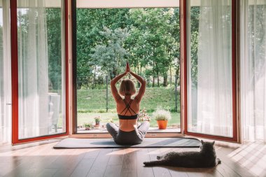 Evde yoga yapan bir kadın, açık pencerenin önünde lotus pozisyonunda oturuyor. Kedisi onu izliyor, yanına uzanıyor..