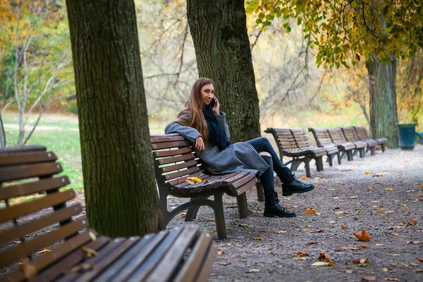 秋の都市公園のベンチに座って携帯電話で話す灰色のコートの若い女性 — ストック写真