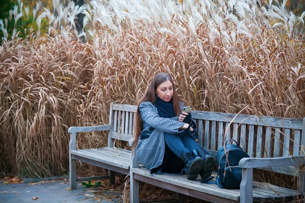 Woman in gray coat sitting on a bench among reeds and searching in her mobile phone