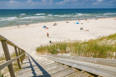 Mavi baltık deniz dalgaları ve güzel, güneşli bir yaz gününün tadını çıkaran turistlerin yer aldığı geniş, beyaz bir kumsal sahiline inen tahta merdiven.