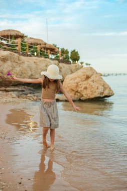 Saman şapkalı bir kızın sakin portresi denizin sığ sularında güzel bir tropikal tatil köyünde duruyor, huzurlu çocukluk anılarını simgeliyor.
