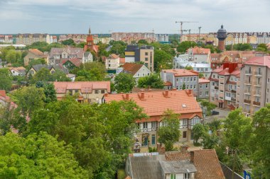 Rus sahil kenti Zelenogradsk 'ın panoramik manzarası, ön planda tarihi kırmızı çatılı binalar ile yoğun yeşillik arasındaki zıtlığı gösteriyor ve arka planda inşaat vinçleri olan modern yüksek binalar