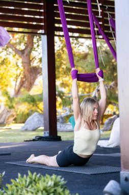 Hava ya da uçuş yogası yapan odaklanmış bir kadın, elinde mor ipek bir hamak tutarken açık hava stüdyosundaki bir paspasın üzerinde poz veriyor.